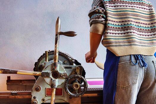 Back view of a printmaker in a patterned sweater and apron operates a traditional mechanical printing press. Artist during the printmaking process photo