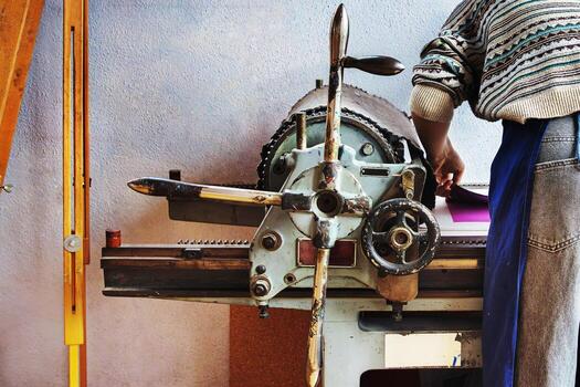 Printmaker in a patterned sweater and apron operates a traditional mechanical printing press. Back view of the artist during the printmaking process photo