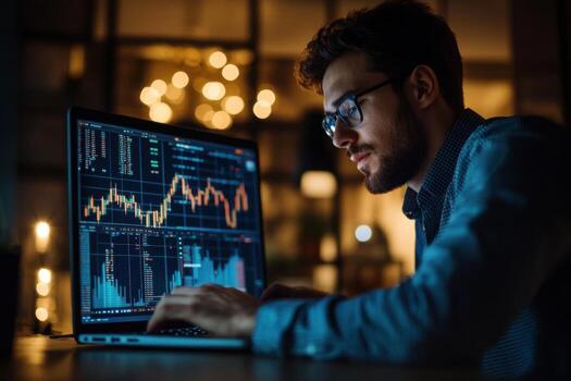 A man in glasses is looking at a laptop screen with stock market graphs photo