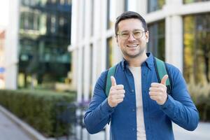 un sonriente hombre vistiendo lentes y un azul camisa da pulgares arriba mientras al aire libre, en pie en un urbano ambiente con edificios en el fondo, exudando confianza, positividad, y un alegre onda. foto