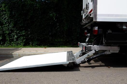 Rear view of delivery truck with hydraulic loading ramp extended onto the street. The ramp is positioned for easy loading and unloading of cargo. The background features greenery and pavement photo