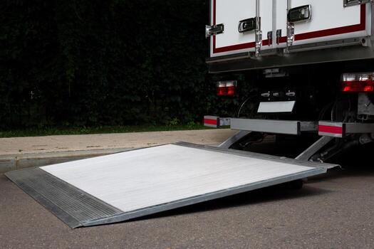 Close-up view of a truck with an unfolded loading ramp on the rear. The ramp is designed for easy loading and unloading of goods. The truck is parked on paved street with green foliage in background photo