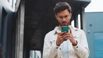sorprendido indio hombre utilizar teléfono inteligente mecanografía hojeada, pierde convirtiéndose sorprendido repentino lotería resultados, malo noticias, fortuna pérdida, fallar al aire libre. árabe hindú chico en céntrico ciudad calle. negocio personas video