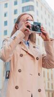 contento caucásico de edad mediana mujer turista de viaje en fines de semana explorador ciudad haciendo foto imágenes en retro Clásico cámara al aire libre. niña en largo Saco en urbano calle durante vacaciones. estilos de vida video