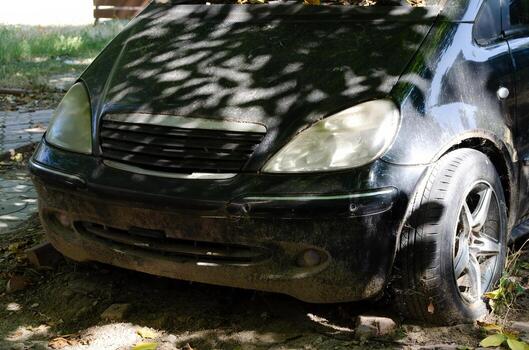 A dirty black car is parked on the side of the road. The car is covered in dirt and has a broken headlight. Scene is one of neglect and abandonment photo