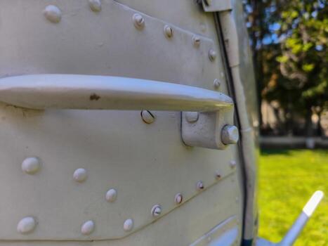 A white metal door with a handle and a bunch of holes. The handle is missing. The door is on a grassy area photo