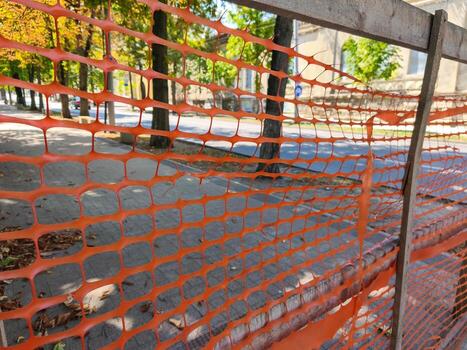 A fence with orange netting is torn and hanging down. The fence is on a sidewalk next to a street photo