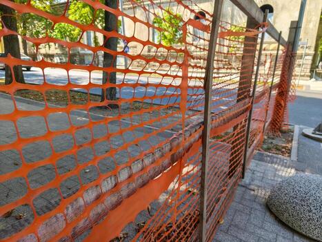 A fence with orange netting on it. The netting is torn and frayed. The fence is on a sidewalk photo