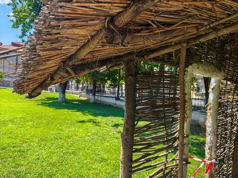 A wooden structure with a thatched roof sits in a grassy field. The structure is surrounded by trees and he is a part of a park photo