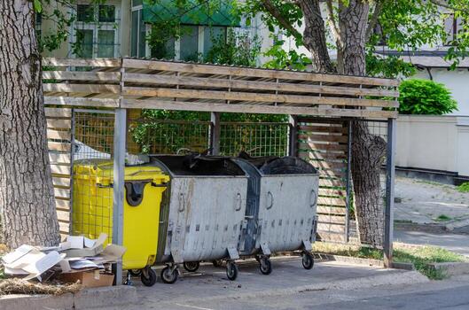 A row of trash cans are lined up under a tree. The trash cans are yellow and gray photo