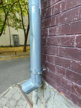 A metal pipe is attached to a brick wall. The pipe is silver and has a black band around it. The brick wall is brown and has a rough texture. The scene is set in a city, with a tree in the background photo