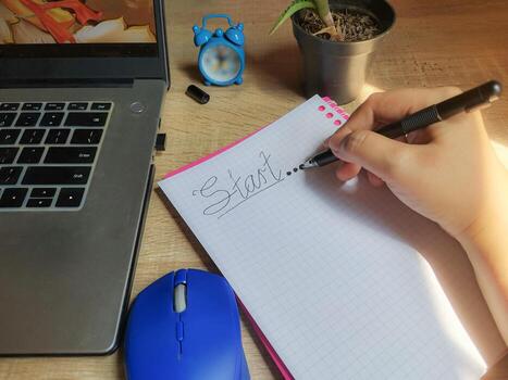 A hand is writing the word Start on a notebook at a wooden desk. This image conveys the beginning of a task, idea, or project, symbolizing productivity, planning, or goal setting. photo