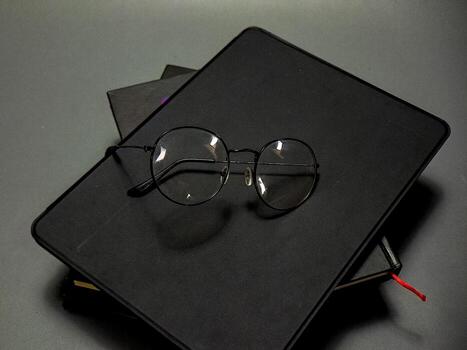 Elegant round spectacles are artfully placed on a dark sketchbook sitting atop more books against a muted grey backdrop, creating a sophisticated and scholarly visual statement. photo