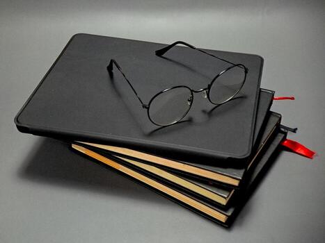 A stack of dark-covered notebooks and a thin pair of glasses rest on a gray surface, evoking a sense of calm focus, study, and the quiet pursuit of knowledge or organization. photo