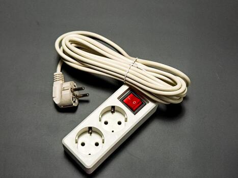 A white power strip with two sockets, a red switch, and a coiled power cord and plug are displayed on a plain grey surface, portraying a simple and functional setting. photo