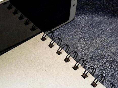 A spiral notebook with black rings sits partially under a device, placed atop a textured surface, highlighting the simple tools of organization and digital integration. photo