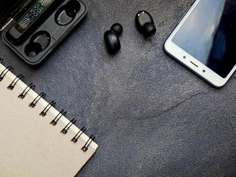 Top-down composition featuring a spiral-bound notebook, cordless earbuds, a charging case and smartphone on a textured dark surface conveys technology and analog elements together. photo