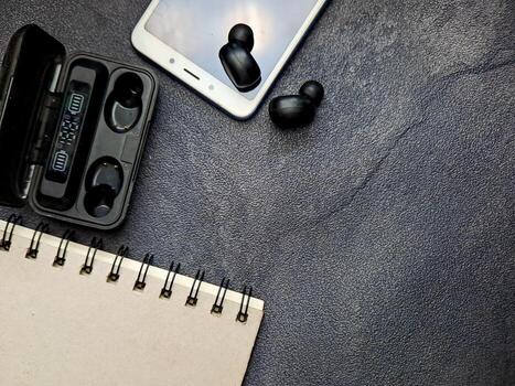 Arrangement features wireless earbuds resting on a smartphone, alongside its charging case and a notepad, set against a textured surface creating a modern, minimalist composition. photo