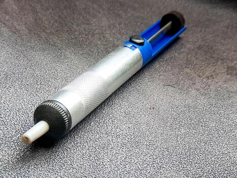 Silver desoldering pump tool showing functional design and utility, with blue accents against a textured surface, showcasing precision and practical craftsmanship. photo