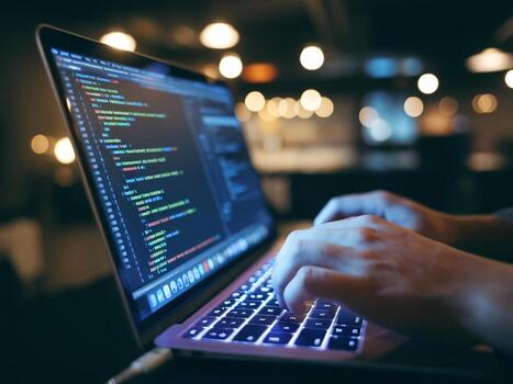 A close-up of a person coding on a laptop in a dimly lit workspace, with colorful code on the screen. photo
