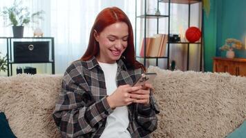 ung Lycklig kvinna på Hem textning på smartphone med fokus och skrattande medan chattar i budbärare app. caucasian rödhårig flicka på soffa njuter roligt konversation, reagerar med glädje till rolig meddelanden video