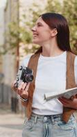 caucásico joven mujer turista de viaje en fines de semana explorador pueblo mirando ciudad mapa, haciendo foto imágenes en retro Clásico cámara al aire libre. contento adulto dama viajero en urbano ciudad calle. estilos de vida video
