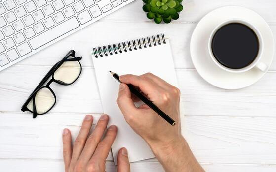Man's hands write in an empty notebook on the office desktop. Workplace with notepad, keyboard, glasses and coffee. Top view. Flatlay. photo