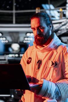 Spaceship engineer using laptop to fix unknown hull breach causing alarm to sound. Space station technician reacts to system failure as radiation spike triggers emergency lights inside orbital cockpit photo