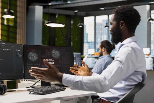 Software developer in office surprised by critical error warning popup, struggling to fix it. African american man troubleshooting error notification on PC screen interrupting workflow photo