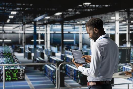 African american admin analyzing infrastructure performance outputs on tablet while he stands on industrial platform. Server room employee finetuning mainframes configurations. photo