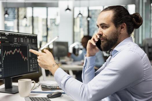 Trader following stock exchange trends on trading platform, talking over phone call with customer. Financial advisor making trading decisions while talking on smartphone with client at computer desk photo