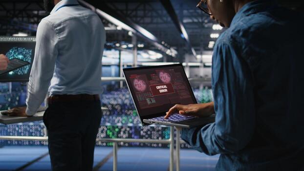 Technician in data center stressed by critical error popup on laptop during diagnostics session. Anxious IT specialist troubleshooting system fault affecting infrastructure performance, photo