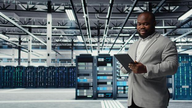 Cheerful data center programmer looking at code debugging tools running on tablet. Server farm IT professional typing on device touchscreen, writing script automating tasks, camera A photo