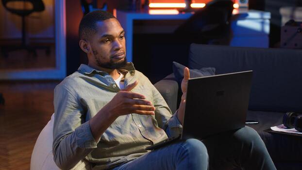 Black man joining online lesson to work on new skills from home space, attend web conference call and watching lecture from loft. Person participates at a virtual training class. Camera B. photo