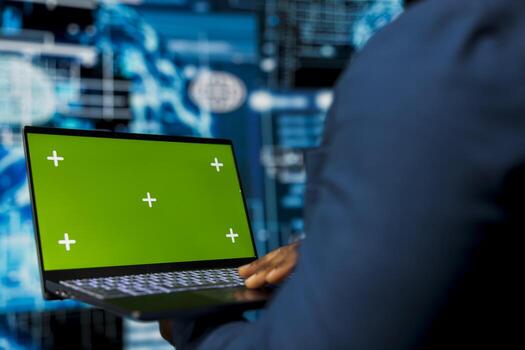 Young male specialist reviews algorithmic data next to green screen, uses laptop surrounded by digital visuals. AI smart technology and neural networks in a high tech environment. photo