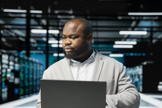 Data center admin using laptop, preparing recovery backups, doing checkup to prolong hardware life span. Server hub employee monitoring infrastructure rigs, auditing systems for vulnerabilities photo