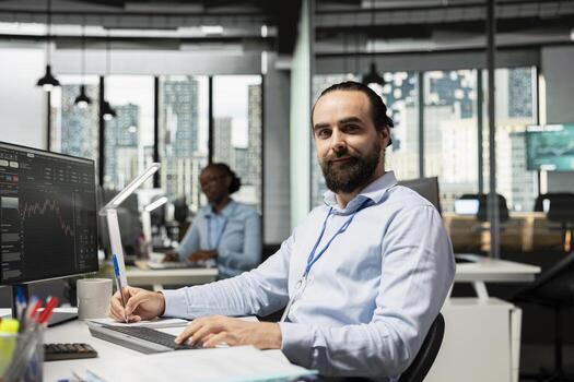 Portrait of smiling trader taking notes while analyzing candlestick charts and price graphs on screen. Cheerful proprietary firm employee writing analysis of market trends and trading strategies photo