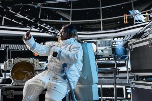 Smiling astronaut doing remote videocall with command center on Earth using smartphone. Jolly African american woman in orbit station chatting with ground control researchers using phone photo