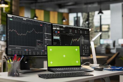 Green screen laptop in brokerage office next to workstation monitor displaying live stock charts. Mockup notebook on desk and computer with financial software in prop firm workspace photo