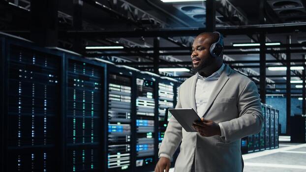 Data center admin wearing headphones using tablet to identify bottlenecks. Server farm IT expert listening music while troubleshooting bugs, implementing optimizations, camera A photo