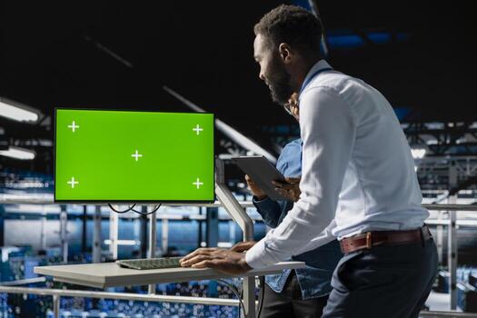 African american IT experts using chroma key device to analyze neural network systems. Data center admin uses green screen pc next to colleague, reviewing AI visualization in background. photo