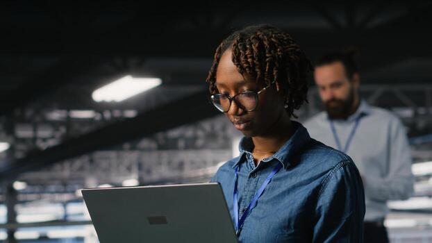 System administrators on data center industrial platform analyzing performance outputs on laptop. Server hub teamworking colleagues finetuning server rig configurations using notebook photo