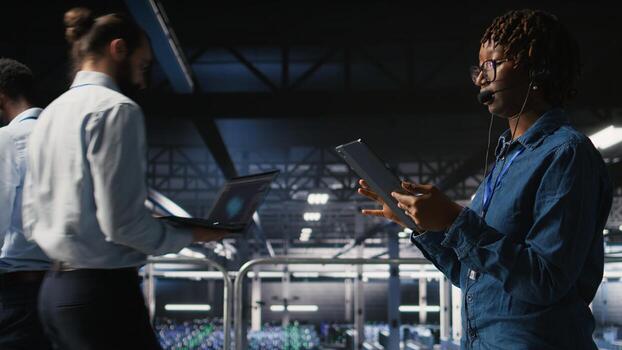 Server room admin using tablet and headset microphone to provide infrastructure tech support. Data center IT specialist resolves backend issues while coordinating with remote teams, photo