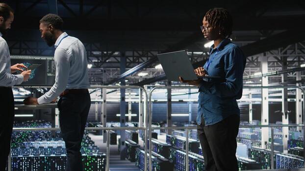 Data center system administrators doing maintenance tasks on laptop, ensuring optimal performance. IT specialists cooperating to review diagnostics and applying patches to server hub gear, photo