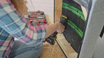 Woman using power drill to screw inner frame of chair, sitting on floor with open toolbox and with dog visible in background video