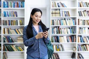 un sonriente joven mujer utilizando su teléfono inteligente en un biblioteca lleno con estantería. ella aparece enfocado y feliz, vistiendo casual ropa, en contra el vívido fondo de pulcramente arreglado libros foto