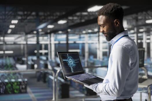 Black male technician in data center using laptop for neural network LLM visualization monitoring. Data scientist on industrial platform overseeing server clusters machine learning processes. photo