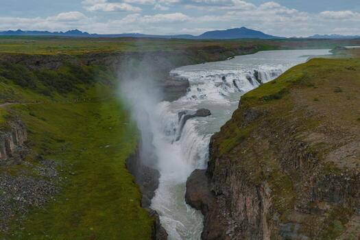Gullfoss waterfall in Iceland flows in two tiers into a canyon, surrounded by green terrain, mist, rolling hills, and distant mountains under a cloudy sky. photo
