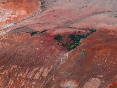 Aerial view of a rugged reddish orange terrain with green vegetation in a triangular depression, showcasing geothermal activity and textured patterns. photo
