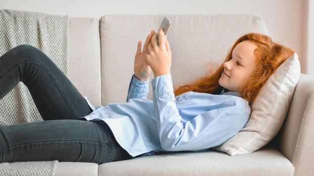 Resting And Relaxing. Portrait of casual generation z girl using her smartphone, lying on the comfortable sofa at home, texting message, chatting with friends on social media network photo